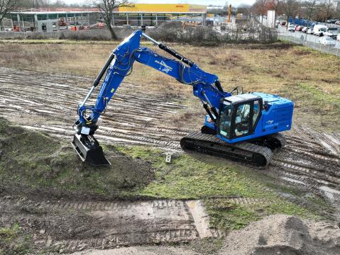 Ein blauer Bagger der Jens Rohde Tiefbau GmbH hebt Erde auf einer Baustelle aus. Im Hintergrund sind Bauarbeiten, ein DHL-Gebäude und eine Straße mit parkenden Autos zu sehen.