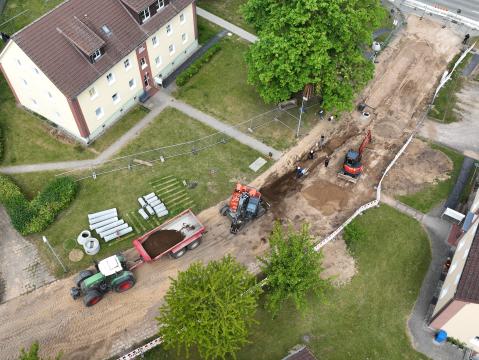 Luftaufnahme einer Baustelle in einem Wohngebiet. Mehrere Baufahrzeuge, darunter ein Traktor mit Anhänger und ein Bagger, arbeiten an einer freigelegten Straße. Auf der rechten Seite des Bildes sind mehrstöckige Wohnhäuser und Gehwege zu sehen. Links sind gestapelte Rohre und Materialien auf einer Grünfläche gelagert. Arbeiter sind entlang der Baustelle beschäftigt, und Bauzäune umgeben den Bereich.