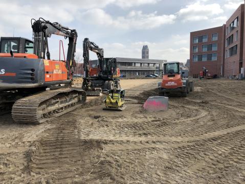 Baustelle mit mehreren orangefarbenen Atlas-Baumaschinen, darunter ein Bagger und eine Walze, auf unbefestigtem Boden. Im Hintergrund sind moderne Backsteingebäude und ein Turm zu sehen. Auf dem Boden liegt ein Betonrohr mit pinker Markierung.