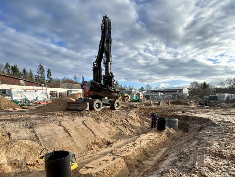 Baustelle mit einem großen Bagger, der auf einem sandigen Gelände steht. Mehrere große, schwarze Rohrstrukturen sind in den Boden eingelassen. Ein Arbeiter befindet sich neben einer der Rohrstrukturen. Im Hintergrund sind Baucontainer, weiteres Baumaterial und Gebäude zu sehen, während der Himmel teilweise bewölkt ist.