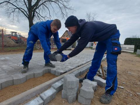 Zwei Arbeiter in blauer Arbeitskleidung verlegen gemeinsam Pflastersteine auf einem Gehweg.