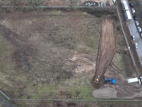 Luftaufnahme einer Fläche mit Erdarbeiten. Ein gelber Bagger und ein blauer LKW befinden sich am rechten Rand der Baustelle. Umgeben ist die Fläche von Bäumen und einem angrenzenden Parkplatz mit geparkten Fahrzeugen.