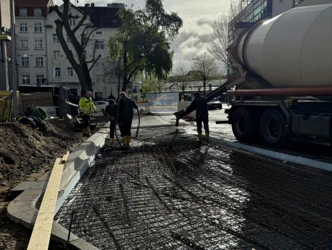 Nahaufnahme eines Straßenbauprojekts mit zwei Arbeitern, die Beton auf einer Bewehrung verteilen. Eine Betonmischmaschine ist im Hintergrund, während im Vordergrund ein halb fertiggestellter Gehweg zu sehen ist.