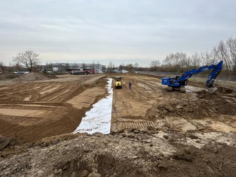 Baustelle mit zwei Baumaschinen, darunter ein blauer Bagger und eine gelbe Planierraupe. Der Boden ist frisch aufgeschüttet und teils mit einer weißen Folie bedeckt. Im Hintergrund sind Bäume und Industriegebäude sichtbar.