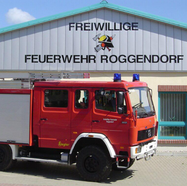 Rotes Feuerwehrfahrzeug mit Blaulicht, geparkt vor einem Gebäude mit der Aufschrift "Freiwillige Feuerwehr Roggendorf". Das Gebäude hat eine weiß-graue Fassade mit einem Logo eines Feuerwehrhelms. Im Hintergrund ist ein blaues Fenster und ein Teil des Himmels zu sehen.