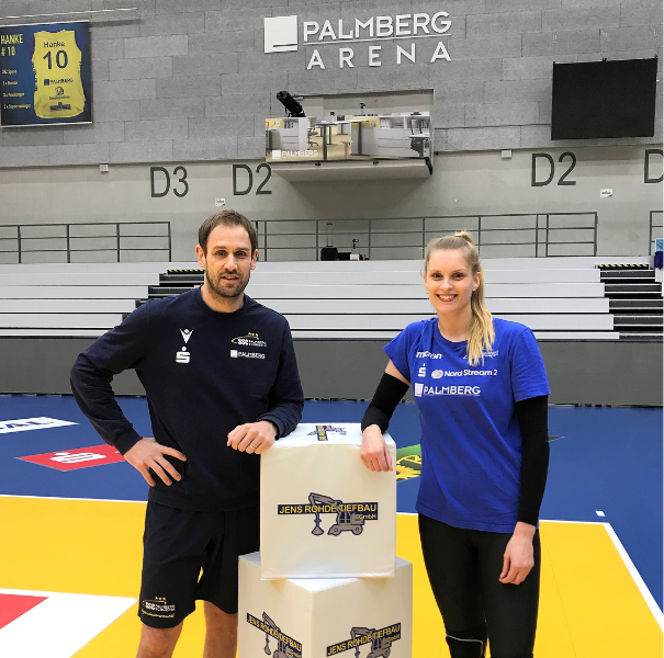 Zwei Personen in Sportkleidung stehen in der Palmberg Arena vor einem Volleyballfeld. Links ein Mann in dunklem Trainingsanzug mit dem Schriftzug „Palmberg“ und rechts eine Frau im blauen Shirt mit Logos von „Nord Stream 2“ und „Palmberg“. Vor ihnen stehen gestapelte Würfel mit dem Logo von „Jens Rohde Tiefbau“. Im Hintergrund sind leere Tribünen und die Beschriftung „Palmberg Arena“ an der Wand sichtbar.